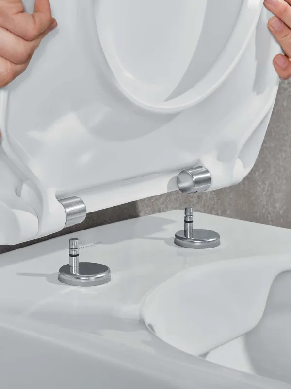 Attaching a Villeroy & Boch toilet seat A person attaching a white toilet seat to a toilet bowl using two metal screws and hinges.