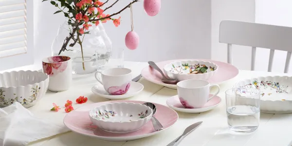 Un tavolo da pranzo luminoso e primaverile, apparecchiato con stoviglie dai colori pastello delle collezioni Rose Garden e Spring Fantasy, decorato con motivi floreali e uova di Pasqua appese a un ramo come centrotavola.