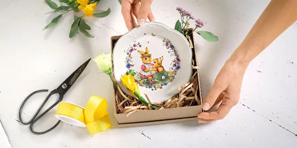 Des mains déposent une assiette décorative Spring Awakeining avec un motif de lapin dans une boîte de paille, entourée de ciseaux, d'un ruban jaune et de fleurs.