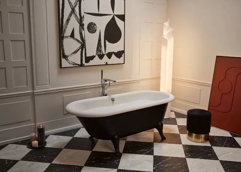 Une salle de bains élégante avec une baignoire en îlot Hommage noire et blanche, un carrelage de sol à carreaux, un tabouret foncé avec un pied doré, des bougies et une grande œuvre d’art géométrique.