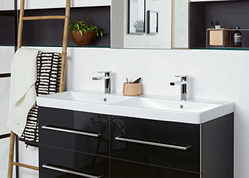Une salle de bains moderne avec un double lavabo Avento avec des tiroirs noirs, un grand miroir, une échelle en bois avec une serviette blanche, une plante et une horloge au mur.