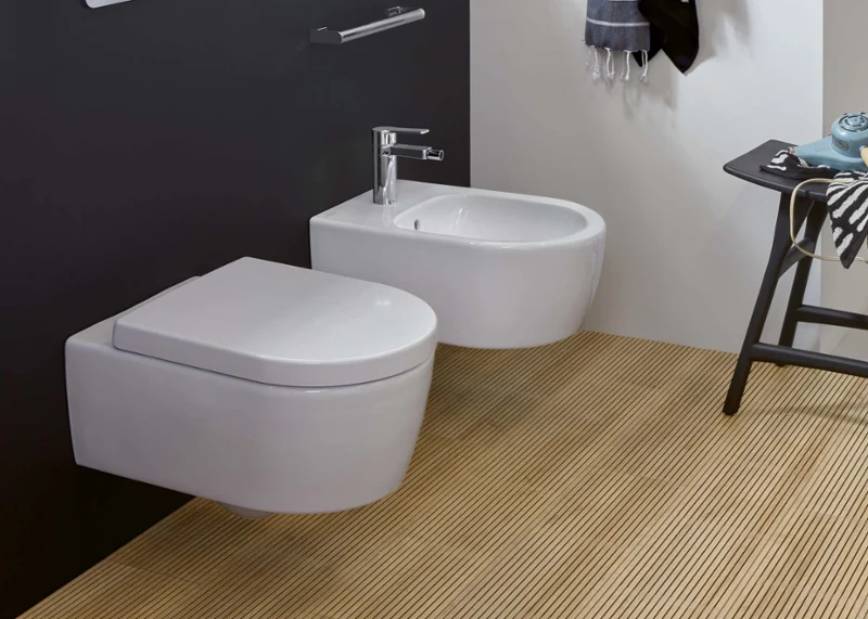 Une salle de bains moderne avec des toilettes et un bidet suspendus de la collection Avento, une serviette sur un crochet, une petite plante sur une étagère et une horloge. Une fenêtre permet à la lumière naturelle de pénétrer.
