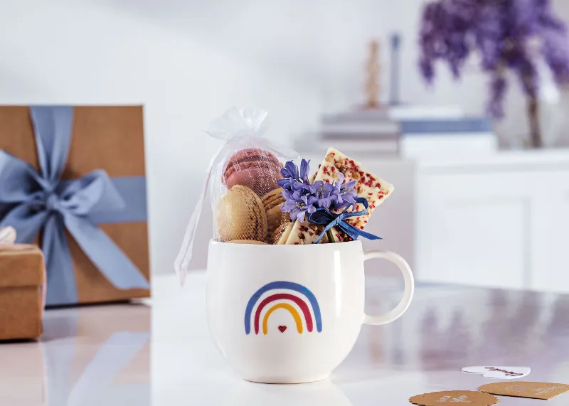 Une tasse « With Love » de Villeroy & Boch au design arc-en-ciel, remplie de macarons, d’une tablette de chocolat et avec fleurs violettes sur une surface blanche avec des boîtes cadeaux et des livres en arrière-plan.