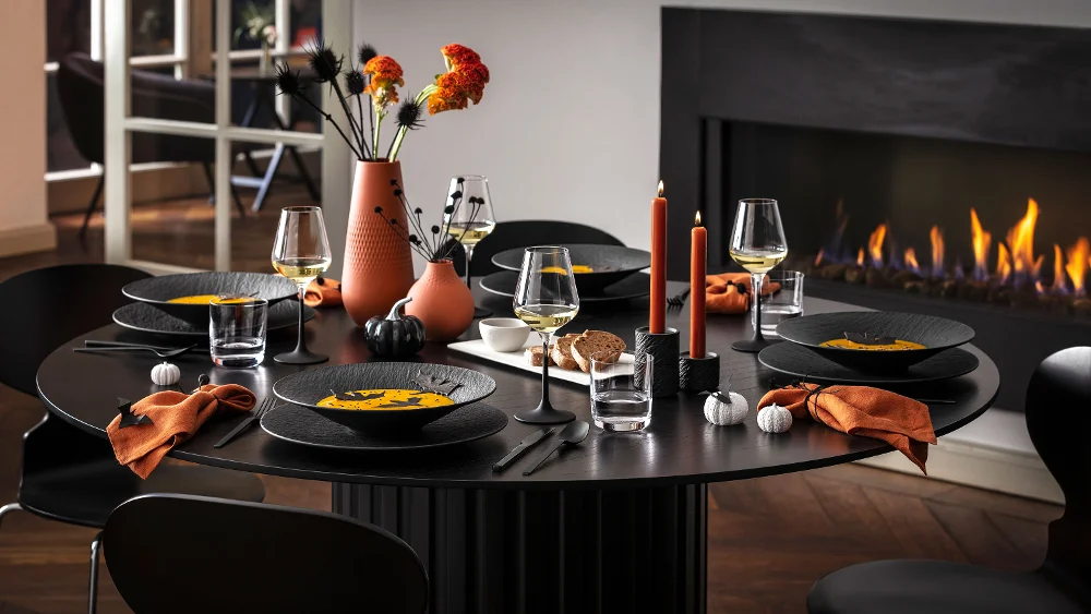 A modern dining table for five with black Manufacture dinnerware, orange napkins, and autumnal decorations, including vases of flowers and lit candles, next to a burning fireplace.