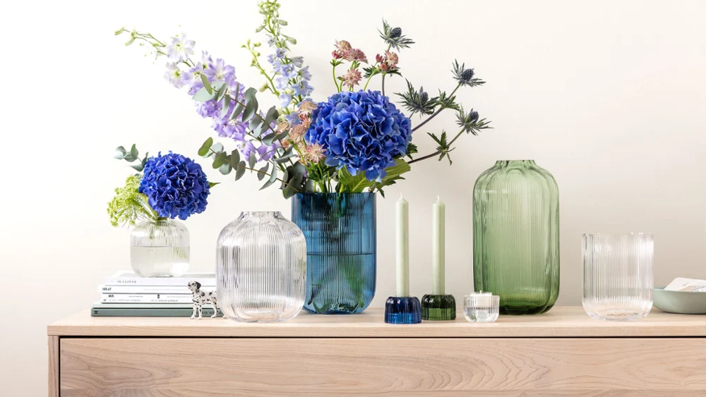 Un sideboard avec des vases Fleur de Villeroy & Boch Un sideboard avec des vases Fleur de Villeroy & Boch, ornés de fleurs bleues et violettes, à côté de deux bougies vertes et de livres empilés.