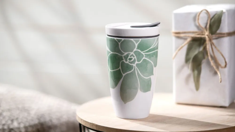 La tazza Coffee ToGo di Villeroy & Boch, una tazza da viaggio in ceramica con un design a foglie verdi, si trova su un tavolo di legno accanto a una confezione regalo legata con uno spago.