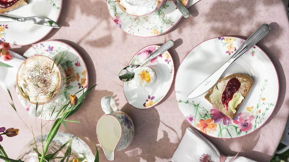 Un set da tavola Mariefleur di Villeroy & Boch con pane tostato e marmellata, un uovo in una tazza e cappuccini. Una lattiera e fiori decorano la tovaglia rosa.