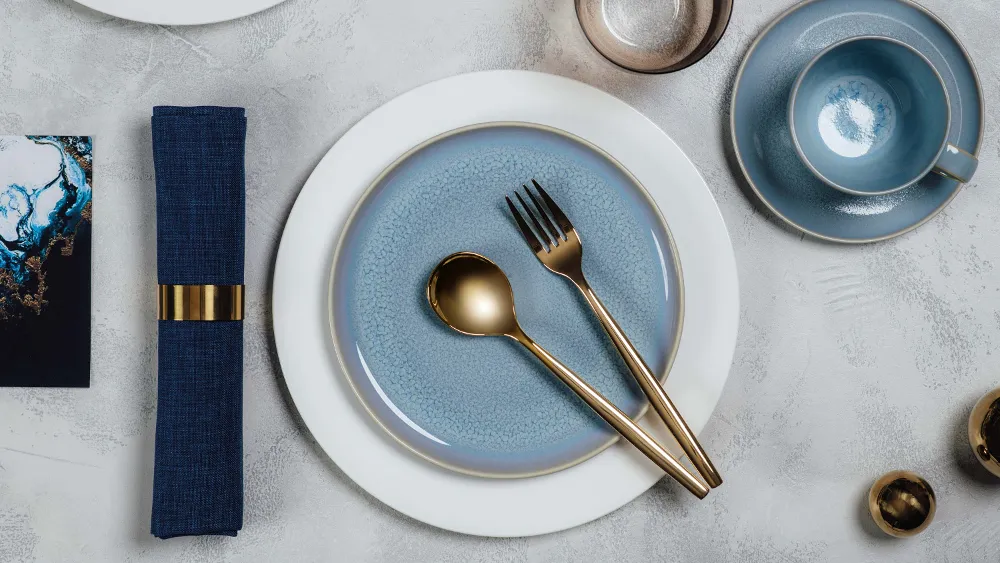 Elegant table decor with blue Crafted dinnerware and white Royal dinnerware, golden MetroChic flatware and a matching napkin.