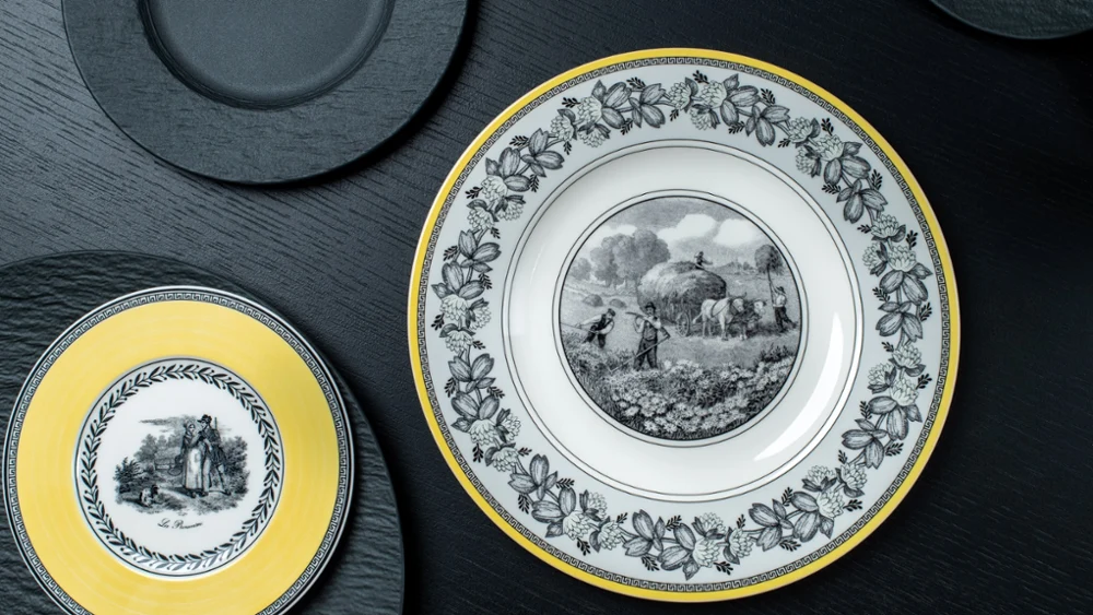 Elegant plates on a dark surface; one large with a rural scene and floral border, another smaller with a yellow rim depicting a couple.