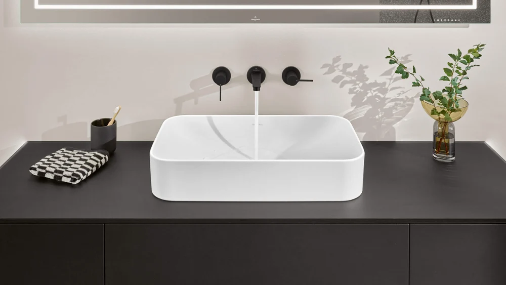 Modern bathroom washbasin with a square design on a black countertop, with a chrome tap and a decorative plant
