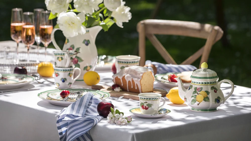 French Garden crockery on an outdoor table Outdoor table setting with floral French Garden porcelain, a teapot, rosé glasses and a cake. White peonies in a vase and fruit are arranged around the table.