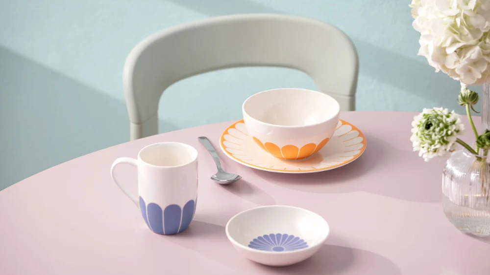 A pastel-coloured table setting with a cup, a bowl and a small plate with blue and orange patterns. Between them is a fork, with a bouquet of white flowers in the background.	