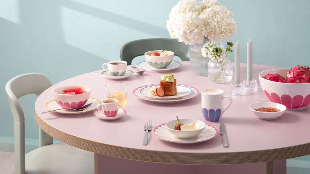 A round table with plates, cups and bowls full of food, decorated with pink patterns. A round table with plates, cups and bowls full of food, decorated with pink patterns. In the centre is a vase of white flowers and two unlit candles. Chairs surround the table.