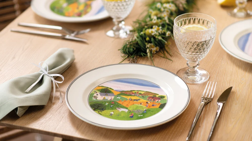 design-naif-placesetting-on-wood-table A dining table set with elegant plates featuring colorful countryside scenes. Crystal glasses with lemon slices and a green floral centerpiece add a fresh touch.