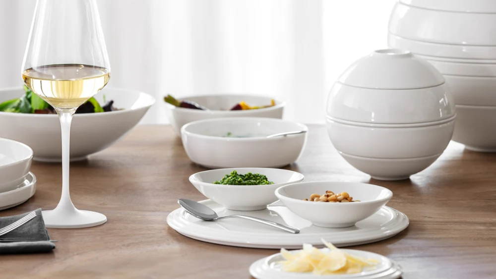 A white wine glass and white ceramic dishes with food are arranged on a wooden table with soft daylight.