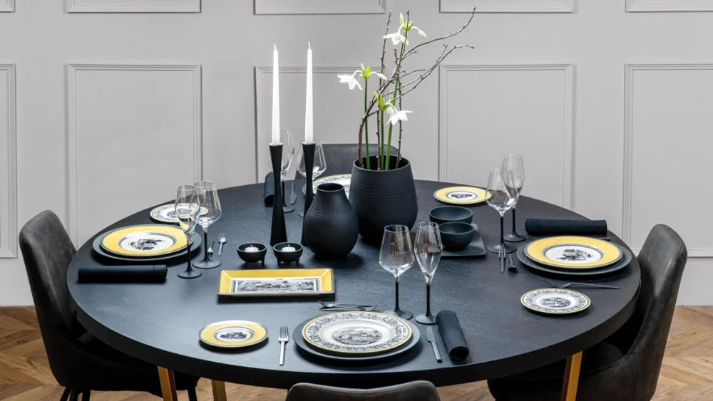 Full table set with Manufacture Rock and Audun Elegant round dining table with black tableware, yellow-rimmed plates, white flowers in a vase, and lit candles, set in a modern room.