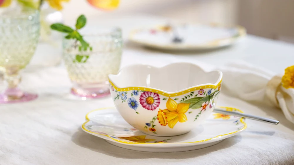 The ‘Spring Awakening’ teacup and saucer with spoon from Villeroy & Boch. The ‘Spring Awakening’ teacup and saucer with spoon from Villeroy & Boch stand on the table next to crystal glasses and softly blurred flowers in the background.