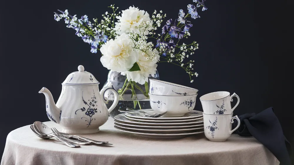 An Old Luxembourg porcelain set with a blue and white floral pattern, consisting of a teapot, cups and plates on a round table, with a vase of white and purple flowers in the background.