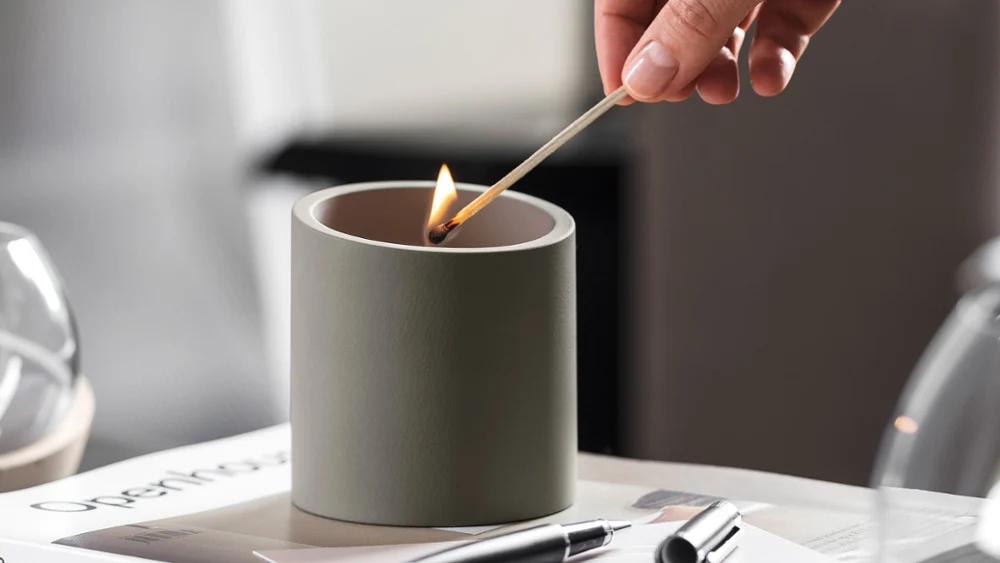 Eine Hand zündet mit dem Streichholz von NewMoon Villeroy & Boch eine Kerze auf einem Tisch an, auf dem Papier und ein Stift liegen.