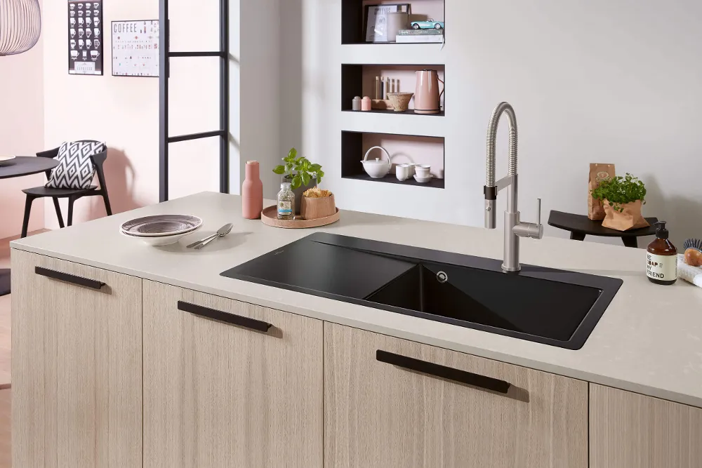 Una cocina moderna con un fregadero negro sobre una isla de cocina de madera clara, un grifo, una planta y varios objetos.