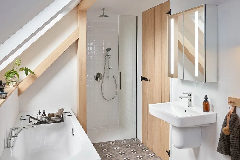 Un cuarto de baño moderno con ducha a ras de suelo, lavabo blanco, armario con espejo y bañera bajo techo inclinado. El suelo de baldosas presenta motivos geométricos. Una puerta y vigas de madera realzan la estancia.