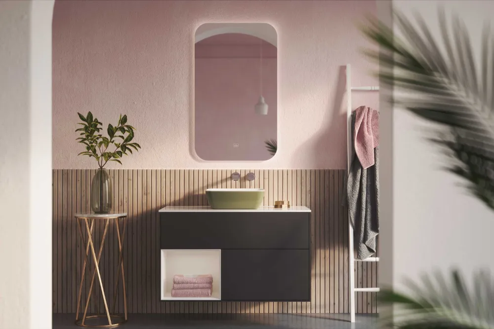 Modernes Badezimmer mit schwarzem Waschtisch, grünem Artis Waschbecken von Villeroy & Boch, rundem Beistelltisch mit Pflanze und rosa Wand mit Spiegel. Handtuchhalter mit Handtüchern auf der rechten Seite und teilweise sichtbarer Palmwedel im Vordergrund.