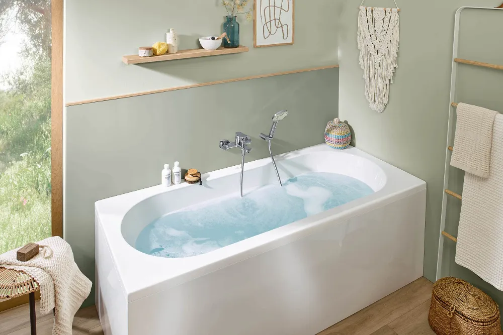 Un cuarto de baño moderno con una bañera llena de agua de la colección O.novo, una estantería de pared con artículos de aseo, un tapiz de macramé y una ventana con vistas a la vegetación.