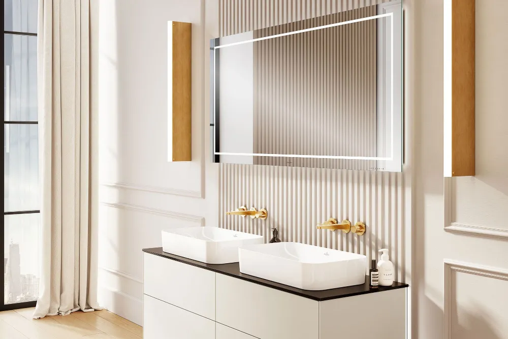 Lavabo doble con grifería dorada de la colección Finion Moderno cuarto de baño con lavabo Finion, grifería dorada en la pared, gran espejo y suelo de madera. La luz del sol entra por un gran ventanal e ilumina la habitación.