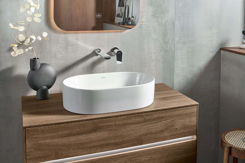 Modernes Badezimmer mit einem weißen ovalen Aufsatzwaschbecken auf einem Holzwaschtisch, einem an der Wand montierten Wasserhahn und einem Fenster mit natürlichem Licht. Zu den dekorativen Elementen gehören eine Vase, ein Hocker und gefaltete Handtücher.