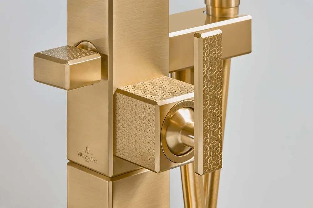 Nahaufnahme einer goldenen Badewannenarmatur mit geometrischem Muster und zwei quadratischen Knöpfen mit strukturiertem Design.
