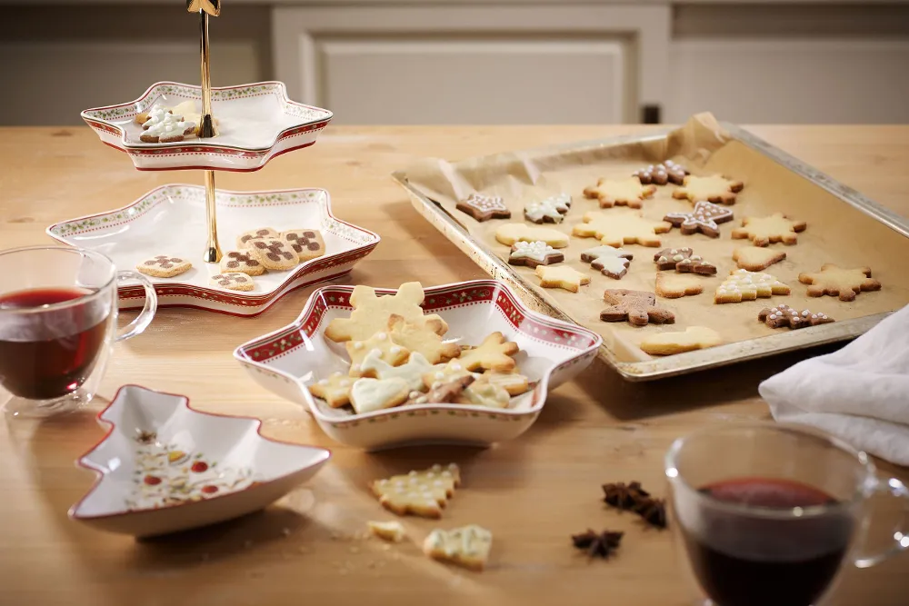 A wooden table with Villeroy & Boch Winter Bakery Delight star-shaped bowls holding decorated biscuits, and a baking tray with festive biscuits on baking paper. There are two glasses with a red drink on the table.