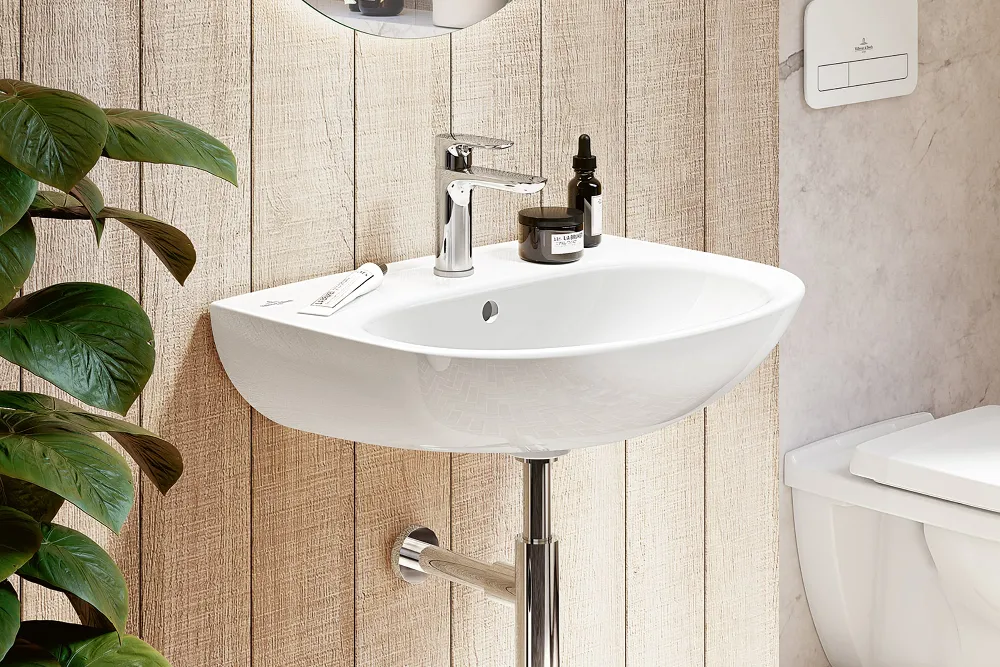 Lavabo afiligranado O.novo de Villeroy & Boch Lavabo con dosificador cilíndrico de jabón y portacepillos de dientes, montado sobre una pared de madera texturizada. A la izquierda se ven plantas verdes y a la derecha parte de un inodoro.