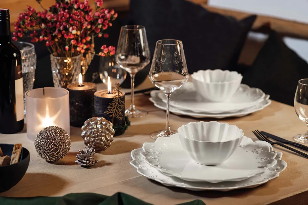 A dining table with white Toy's Delight Royal Classic plates, wine glasses, candles and decorative ornaments. There are also flowers and a bottle of red wine on the table.