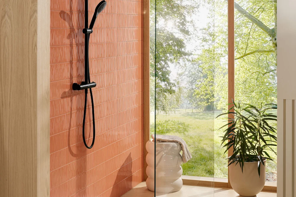 Robinetterie Verve Showers avec receveur Architectura dans une salle de bain lumineuse Douche moderne avec parois en verre et robinetterie noire, à côté d’une grande fenêtre donnant sur la verdure.