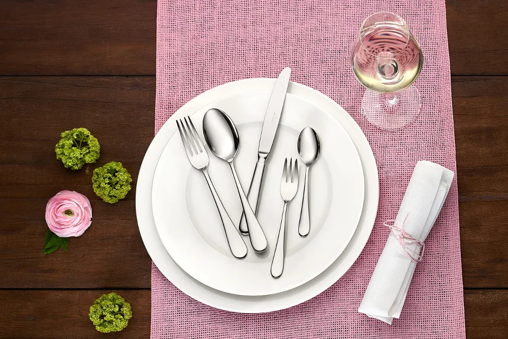The Villeroy & Boch Oscar collection is presented in a carefully arranged table setting. A white plate is complemented by elegant silver cutlery and a wine glass, all arranged on a pink placemat. Next to this is a rolled napkin and bright green and pink flowers.