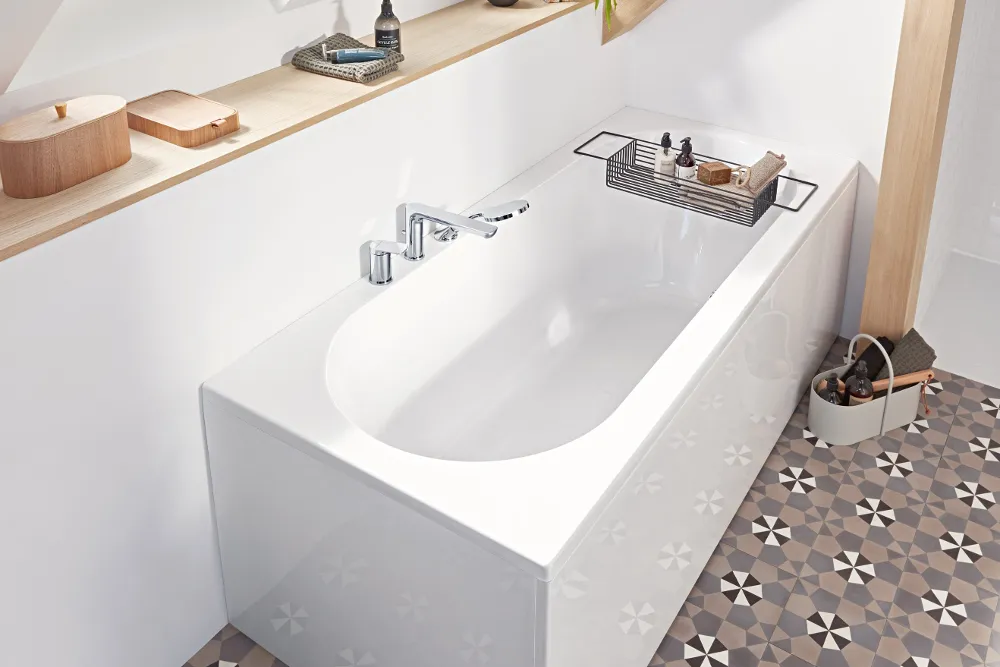 Una bañera blanca de la colección O.novo con grifo plateado junto a una estantería de madera con artículos de aseo. El cuarto de baño tiene azulejos estampados y luz natural que entra por una ventana sobre la bañera.