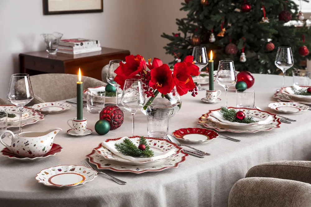 A festive dining table setting with elegant Toy's Delight crockery, lit candles, red flowers in a vase and Christmas decorations, with a decorated Christmas tree in the background.