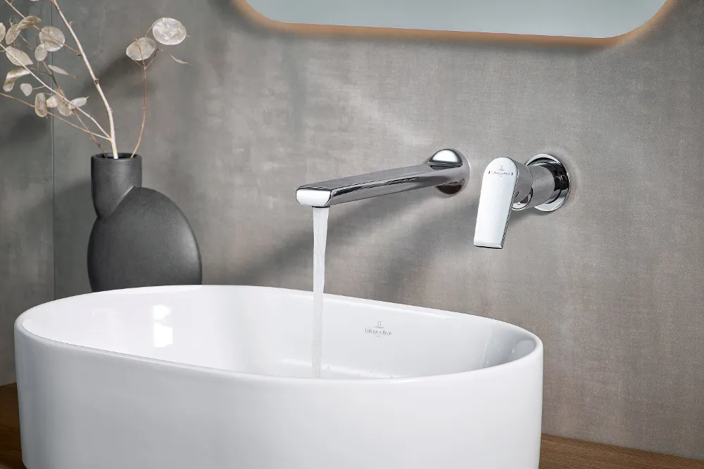 Een moderne wastafel in de badkamer, met water dat uit de verchroomde kraan stroomt. Een zwarte vaas met gedroogde planten staat naast de Villeroy & Boch Collaro wastafel.