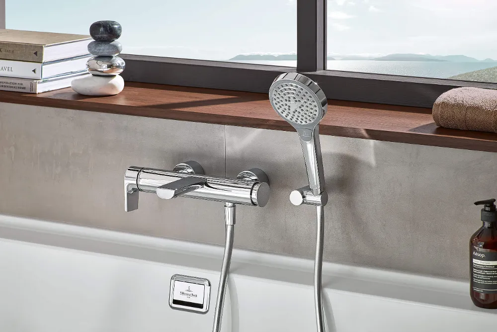 Nahaufnahme einer modernen Badewanne mit verchromtem Handbrausenkopf an der Wand. Auf der Fensterbank darüber liegen Steine. Durch das große Fenster ist eine grüne Landschaft zu sehen.