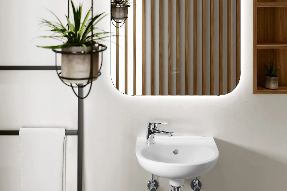 Lavabo O.novo en un baño minimalista con plantas Un cuarto de baño minimalista compuesto por un pequeño lavabo O.novo blanco, un espejo rectangular con listones de madera, una toalla sobre un soporte negro, una pequeña planta en un estante de pared y una planta colgante.