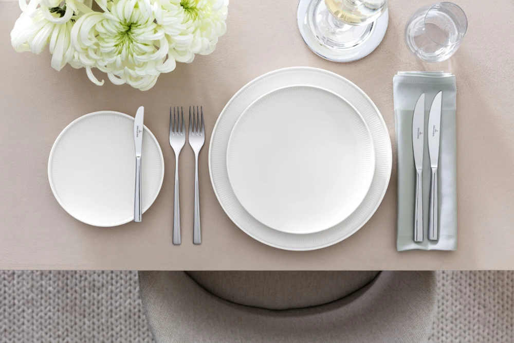 Une table dressée avec de la vaisselle Afina et des couverts de table Piemont Une présentation de table sophistiquée avec la collection Piemont de Villeroy & Boch, comprenant une assiette plate blanche, une assiette à salade, un ensemble de couverts de table, des verres et un centre de table floral sur une nappe beige.