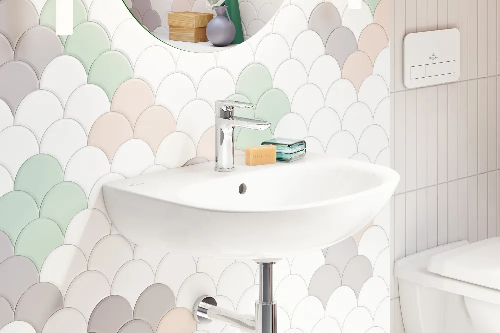 Lavabo O.novo blanco con espejo pequeño Un cuarto de baño moderno con un lavabo O.novo blanco suspendido y un inodoro montado en la pared. La zona del lavabo está equipada con azulejos de colores y un pequeño espejo.
