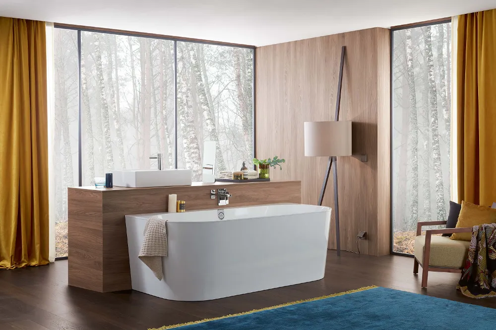 A modern bathroom with a white freestanding bath, wood panelling, a large countertop, full-length windows with yellow curtains, an armchair and a blue rug.