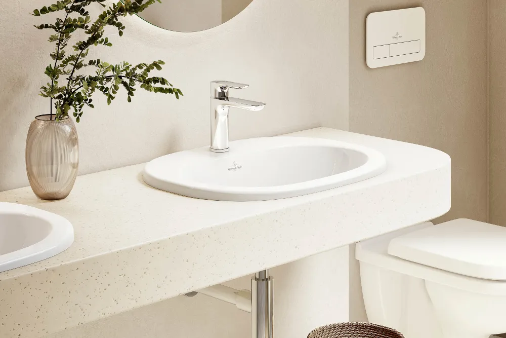 Cuarto de baño con lavabo, espejo e inodoro de la colección O.novo Un cuarto de baño con un lavabo O.novo blanco, un espejo redondo, un grifo y un inodoro montado en la pared. Hay una maceta en la estantería y una cesta de mimbre en el suelo.