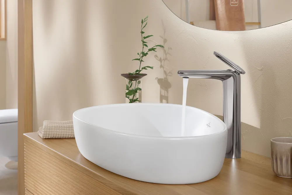Wastafel met een witte ovale wastafel uit de Antao collectie en een elegante, verchroomde kraan met stromend water. Een opgevouwen beige handdoek en een groene plant liggen op de houten plank naast de wastafel.