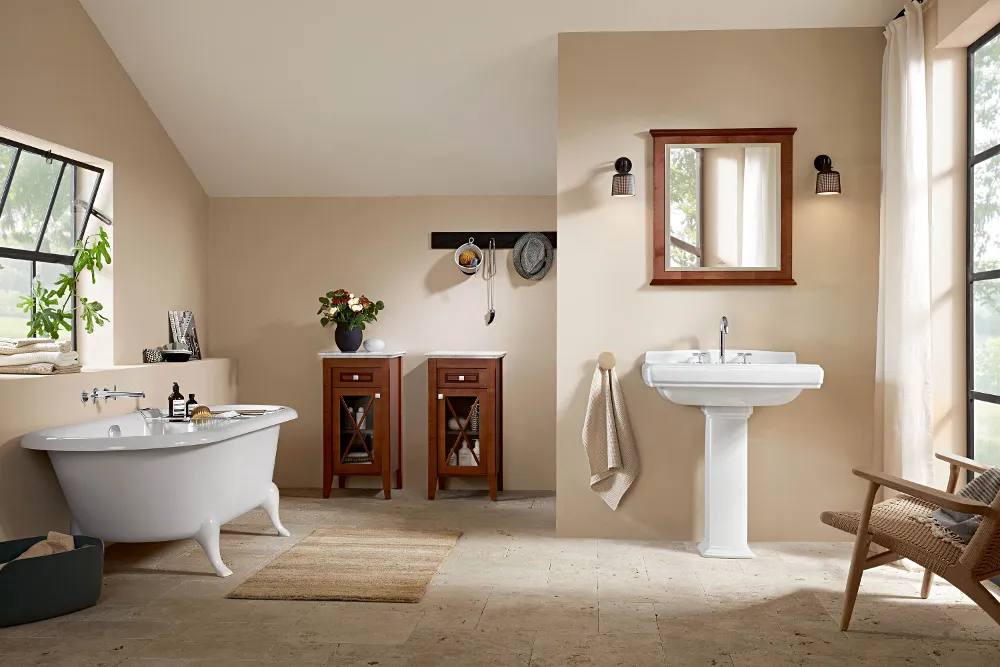 A bathroom with a freestanding bath, a washbasin with a wall mirror and a wooden cabinet with flowers. There are two windows, a chair, a rug and a towel on a hook.