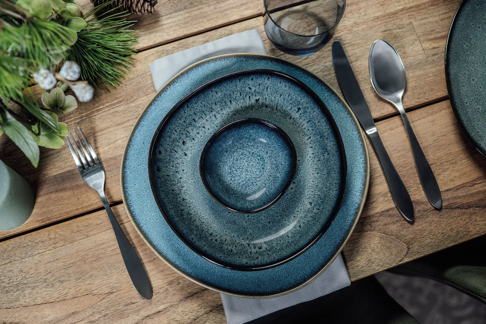 Une décoration de table élégante avec des assiettes Lave et des couverts de table Arthur de Villeroy & Boch sur une table en bois, décorée avec des plantes vertes.