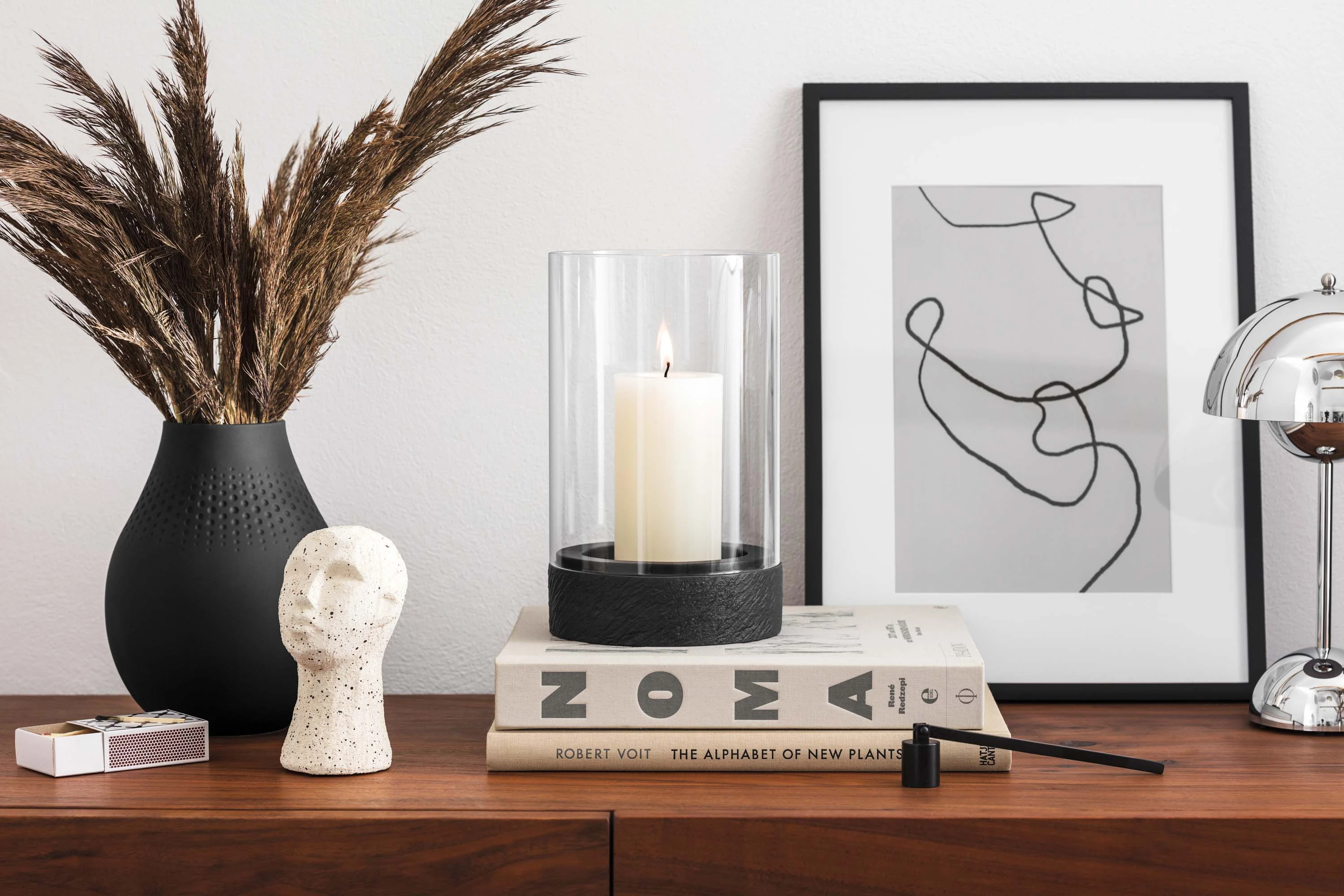 A modern highboard with decorative objects, including a vase with dried plants, a black Manufacture hurricane lamp, a sculpture, stacked books and a piece of framed abstract artwork.