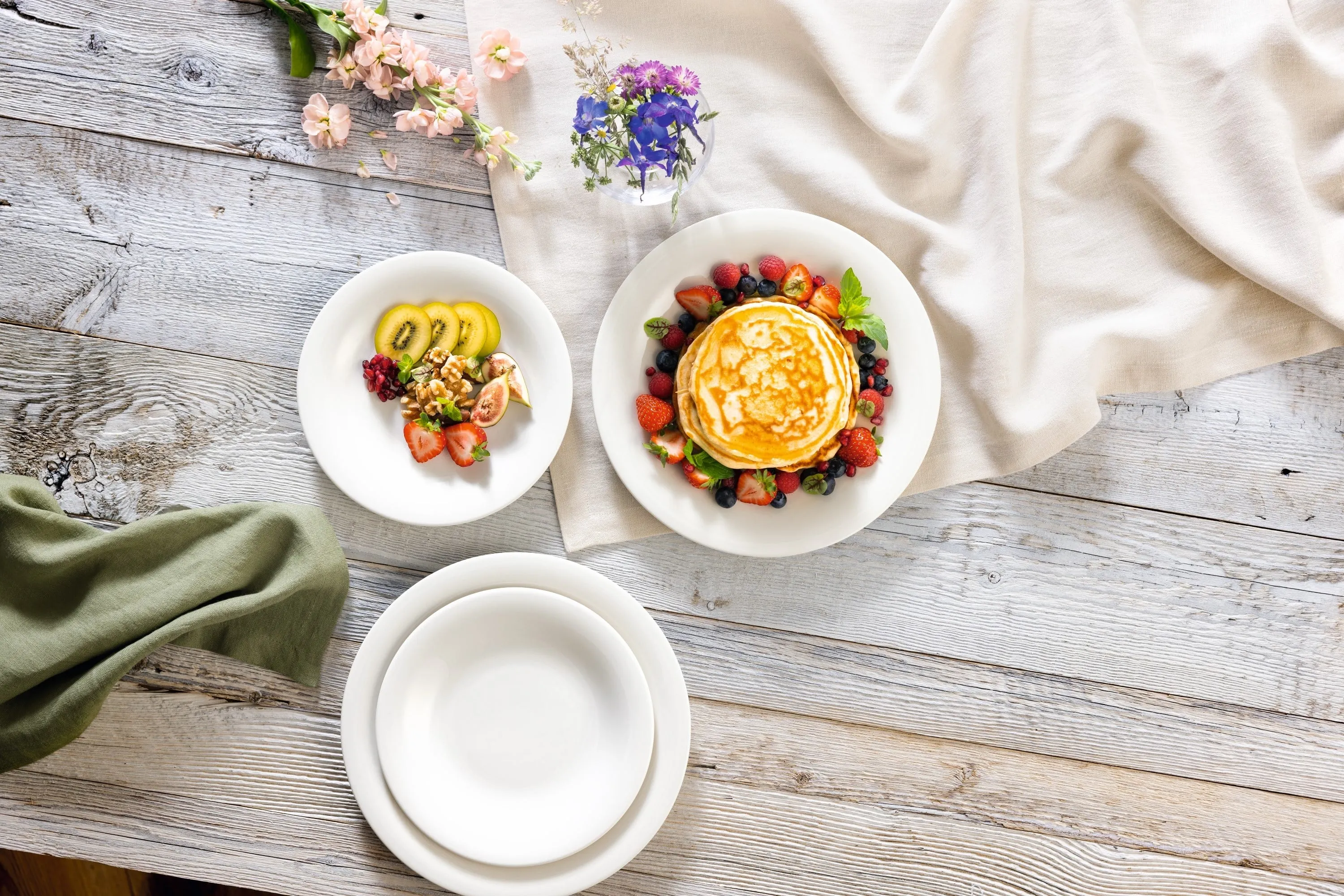 Piatto New Cottage con pancake Una pila di pancake con frutti di bosco viene servita su un piatto New Cottage di Villeroy & Boch, con frutta a fette su un altro piatto. Il tavolo di legno è decorato con fiori e una tovaglia drappeggiata.