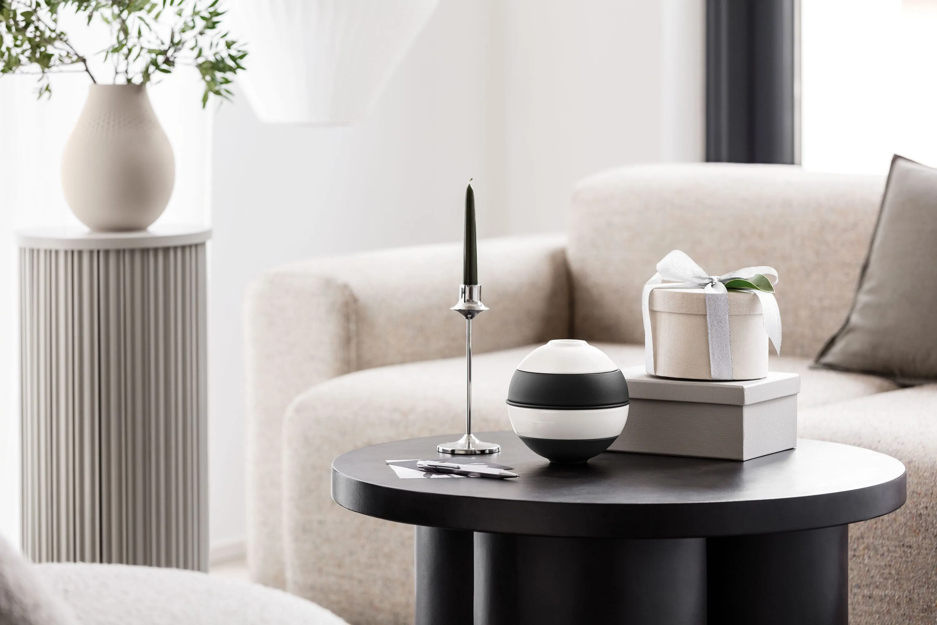 Une table basse ronde noire avec La petite Boule de Villeroy & Boch, une bougie verte sur un support argenté et une boîte cadeau sur deux livres. Un canapé beige et un socle gris avec un vase en arrière-plan.